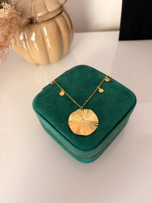 collier chaîne en acier inoxydable doré et orné d'un pendentif rond strié et de petits sequins ronds striés sur les côtés. Bijou présenté sur une boite à bijoux vert émeraude disposée sur une table décorée d'une citrouille en céramique.