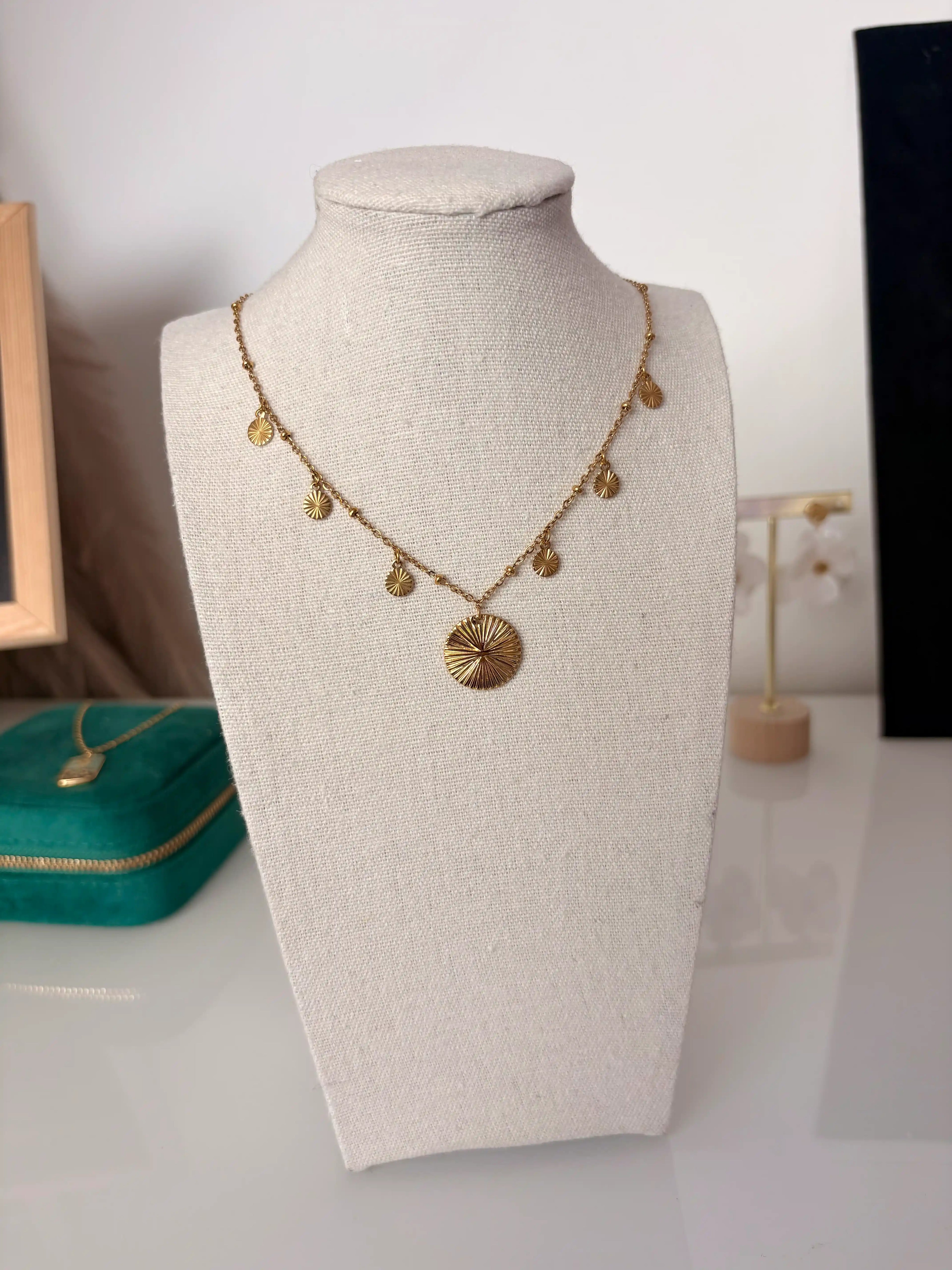 collier chaîne en acier inoxydable doré et orné d'un pendentif rond strié central et de petits sequins ronds striés sur les côtés. Bijou présenté sur un buste en coton beige.
