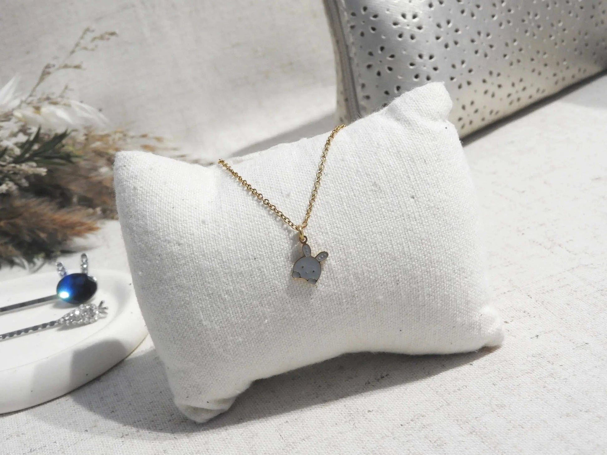 Collier enfant à chaîne fine en acier inoxydable doré et orné d'un pendentif lapin gris en résine époxy et acier inoxydable doré. Bijou posé sur un mini-coussin beige, posé sur une nappe beige décorée d'une trousse et coupe avec des épingles à cheveux.