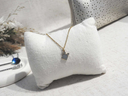 Collier enfant à chaîne fine en acier inoxydable doré et orné d'un pendentif lapin gris en résine époxy et acier inoxydable doré. Bijou posé sur un mini-coussin beige, posé sur une nappe beige décorée d'une trousse et coupe avec des épingles à cheveux.