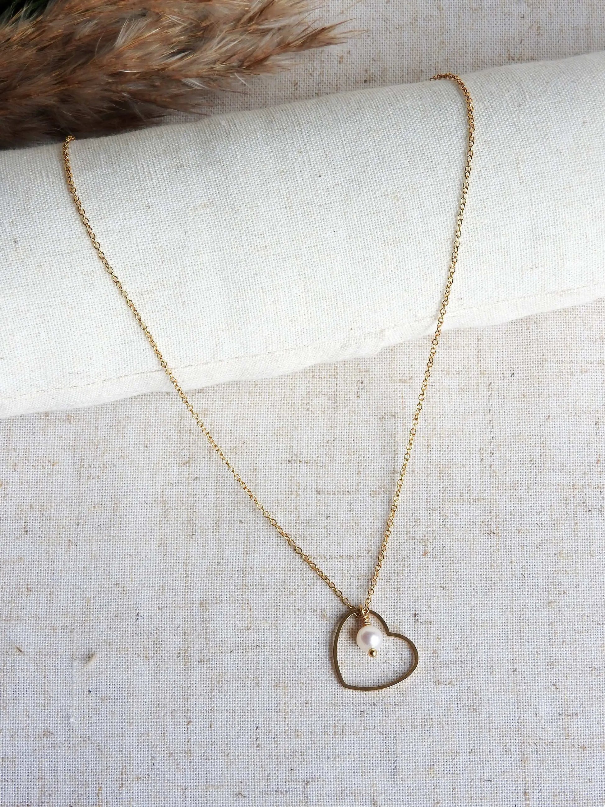 collier chaîne fine avec un pendentif coeur évidé et orné d'une perle d'eau douce. Bijou en acier inoxydable doré posé sur une nappe beige décorée d'une brindille.