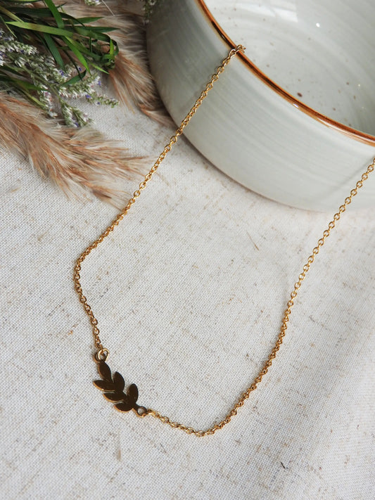 Collier à chaîne fine orné d'une feuille intercalaire en acier inoxydable doré. Bijou posé à plat sur une nappe beige et décorée d'un mini-bouquet de fleurs et un bol en céramique vert pâle.