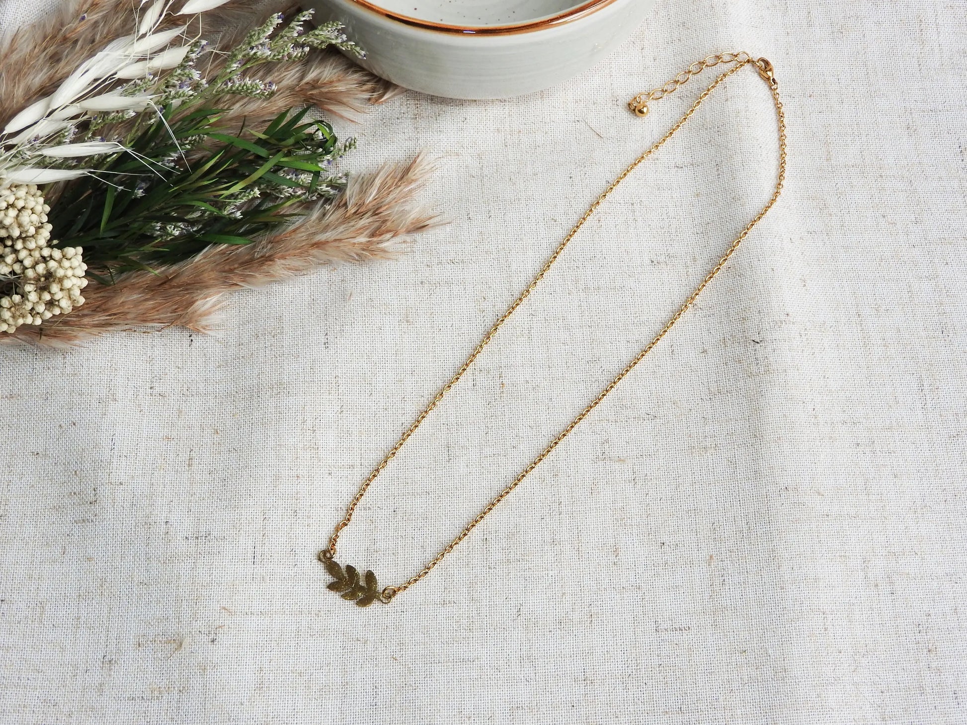 Collier à chaîne fine orné d'une feuille intercalaire en acier inoxydable doré. Bijou posé à plat sur une nappe beige et décorée d'un mini-bouquet de fleurs et un bol en céramique vert pâle.
