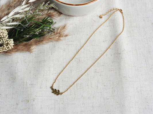 Collier à chaîne fine orné d'une feuille intercalaire en acier inoxydable doré. Bijou posé à plat sur une nappe beige et décorée d'un mini-bouquet de fleurs et un bol en céramique vert pâle.