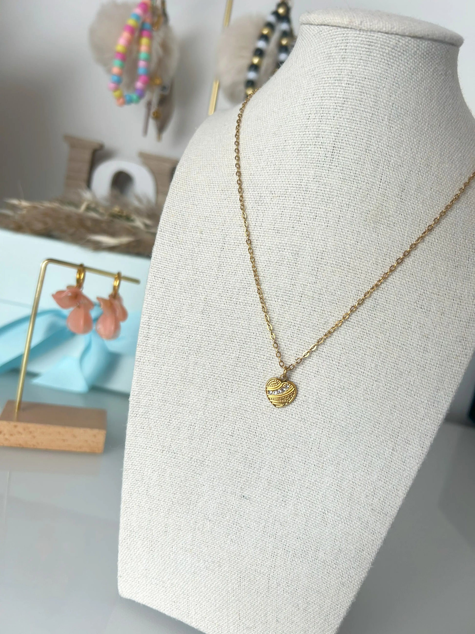 Collier à chaîne fine et pendentif cœur texturé orné de pierres en oxydes de zirconium et en acier inoxydable doré. Bijou présenté sur un buste beige en coton en fond d'image une paire de boucles d'oreilles à pétales couleur pêche et portes-clés colorés. 