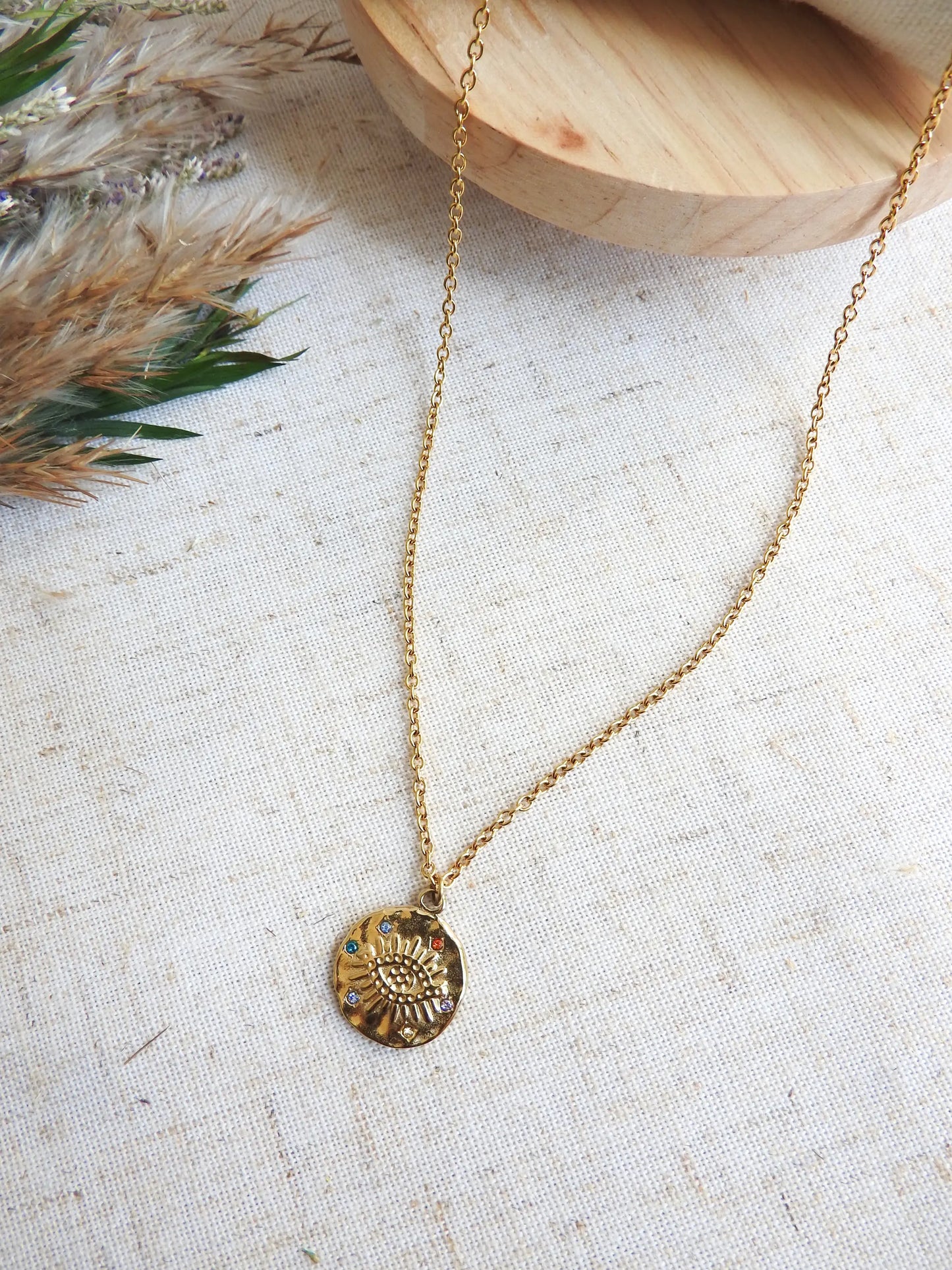 collier chaîne fine en acier inoxydable doré à pendentif médaille décoré d'un oeil protecteur et de strass multicouleurs. Bijou posé à plat sur une nappe décorée d'une brindille et d'un plateau en bois.