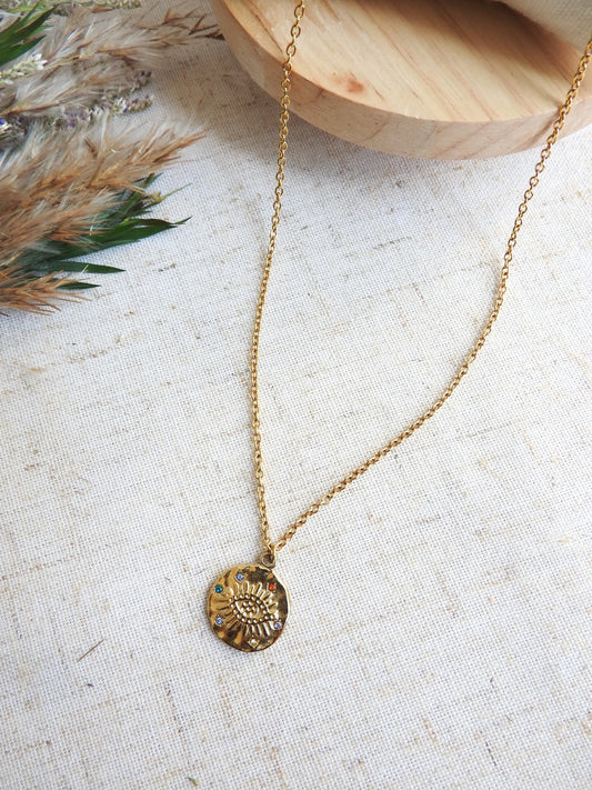collier chaîne fine en acier inoxydable doré à pendentif médaille décoré d'un oeil protecteur et de strass multicouleurs. Bijou posé à plat sur une nappe décorée d'une brindille et d'un plateau en bois.