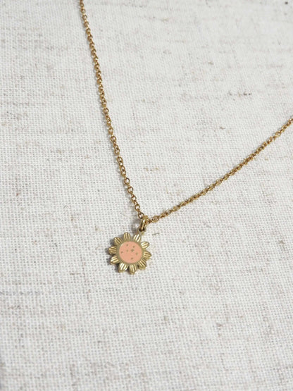 Collier pour enfant à chaîne fine orné d'un pendentif tournesol saumon en résine époxy et acier inoxydable doré. Bijou posé à plat sur une nappe beige en coton.