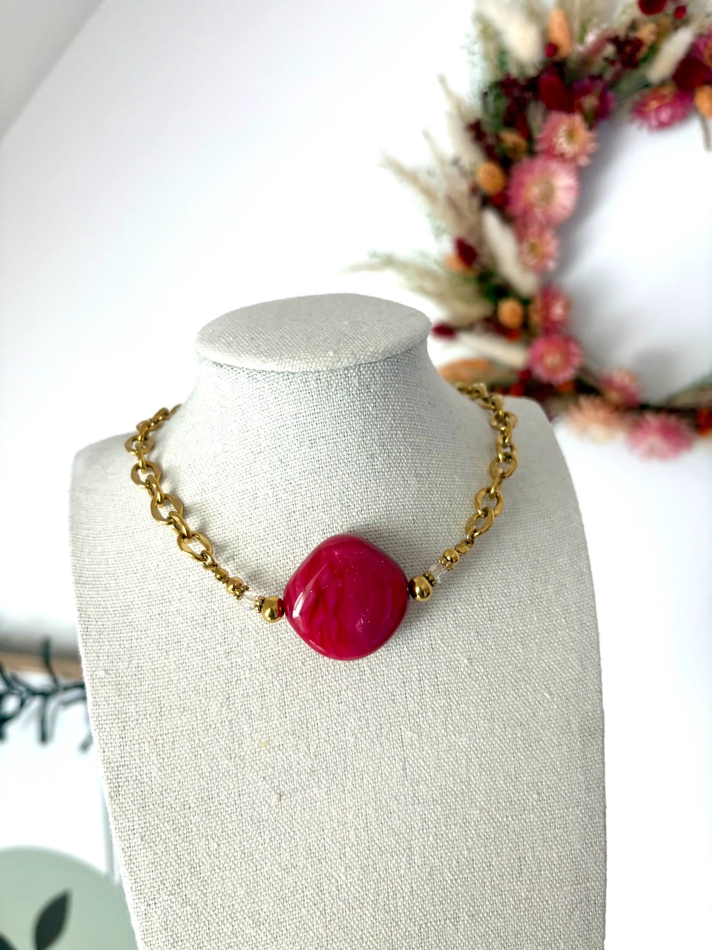 Collier chaîne larges maillons en acier inoxydable doré et perle losange couleur framboise. Bijou posé sur un buste en coton beige sur fond d'une couronne en fleurs séchées.