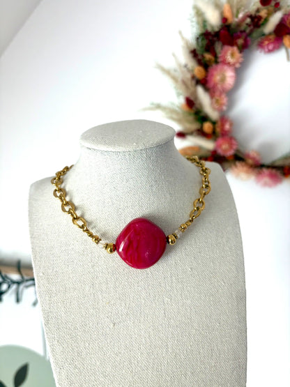 Collier chaîne larges maillons en acier inoxydable doré et perle losange couleur framboise. Bijou posé sur un buste en coton beige sur fond d'une couronne en fleurs séchées.