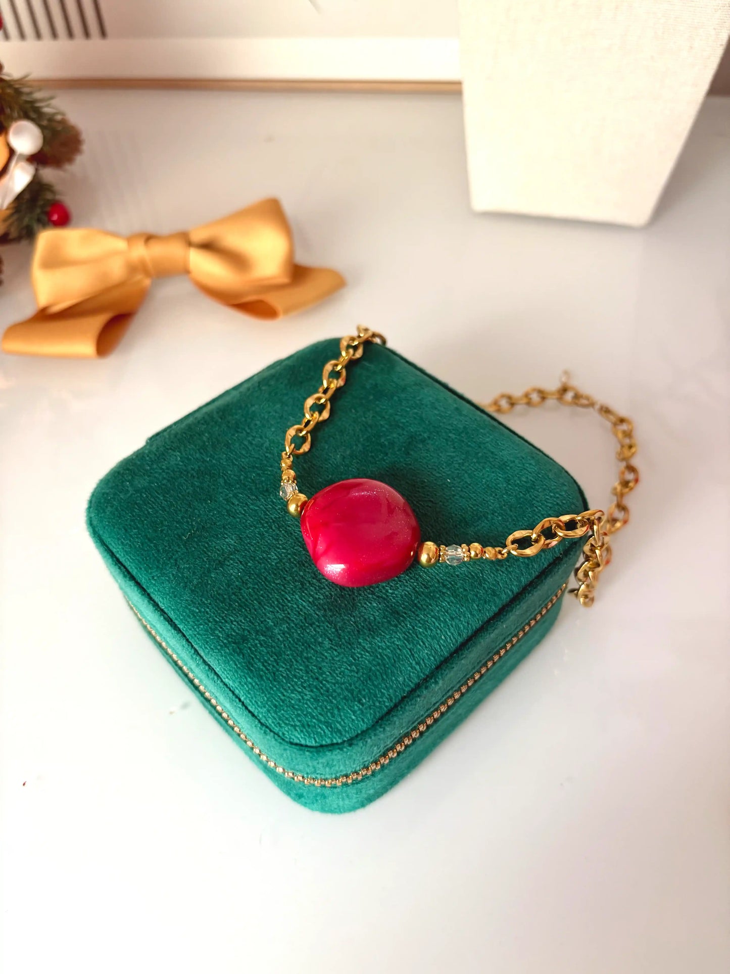 Collier chaîne larges maillons en acier inoxydable doré et perle losange couleur framboise. Bijou posé sur une boite à bijoux vert sapin, posée sur une table décorée d'un nœud moutarde.