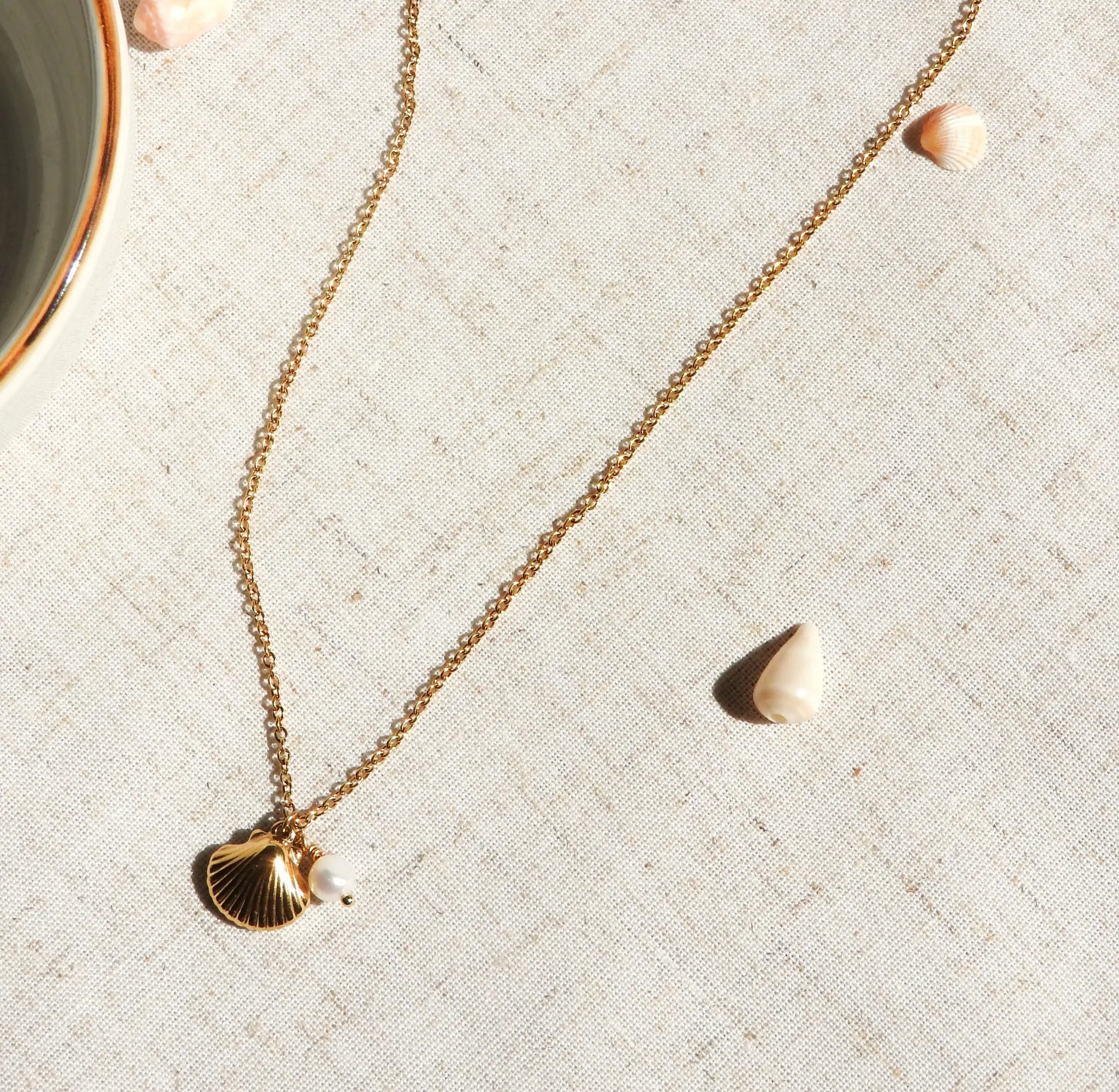 Collier pendentif en acier inoxydable doré, orné d'un pendentif coquillage et une perle d'eau douce blanche. Bijou posé à plat sur une nappe beige décorée de coquillages.