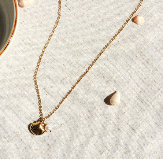 Collier pendentif en acier inoxydable doré, orné d'un pendentif coquillage et une perle d'eau douce blanche. Bijou posé à plat sur une nappe beige décorée de coquillages.