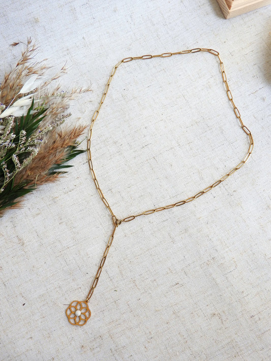 collier chaîne à maille rectangle en acier inoxydable doré posé à plat sur une nappe beige. Bijou orné d'un pendentif fleur filigrane en acier doré, orné d'une pierre de lune. Chainette pendante avec en son bout le pendentif fleur.