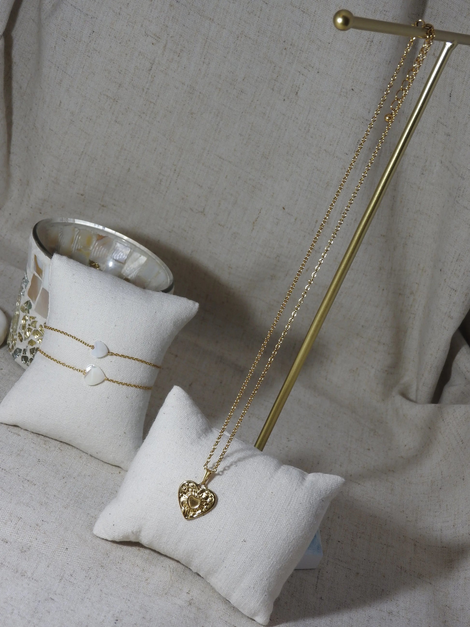 Collier chaîne pendentif coeur gravé de deux mains supportant un coeur. Bijou en acier inoxydable doré présenté sur une barre métaliique doré et un mini-coussin beige en coton. En arrière plan, deux bracelets chaîne avec une perle en forme de coeur en nacre blanche présentés sur un mini-coussin beige.