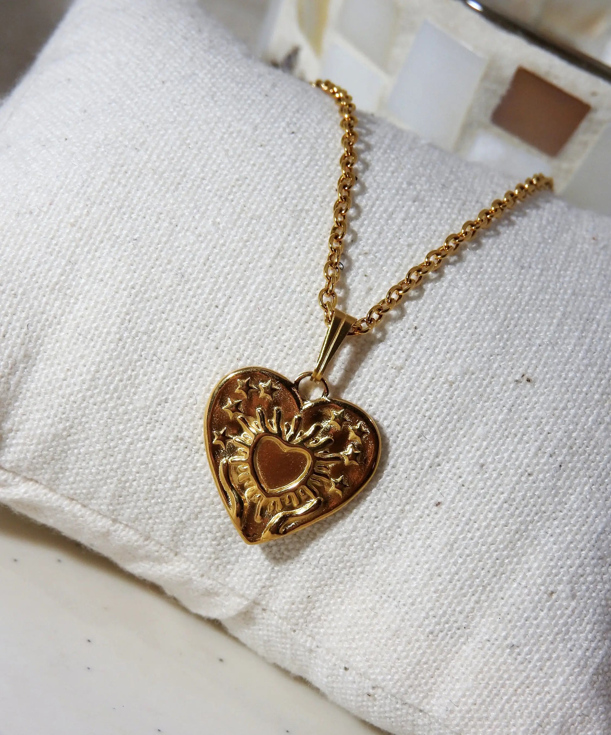 Collier chaîne pendentif coeur gravé de deux mains supportant un coeur. Bijou en acier inoxydable doré posé un mini-coussin beige en coton