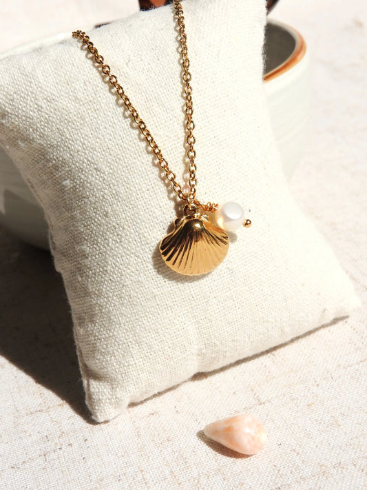 Collier chaine présenté sur un mini-coussin beige en coton. Bijou orné d'un pendentif coquillage en acier inoxydable doré et une perle d'eau douce blanche.
