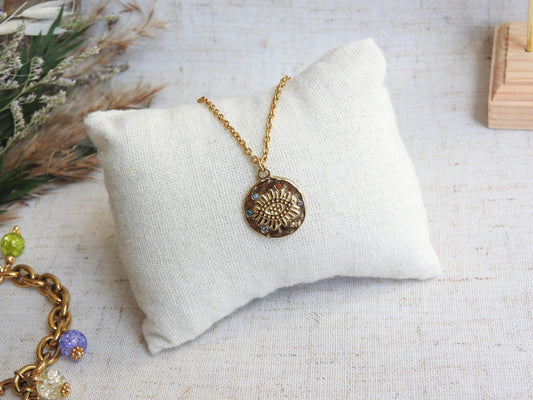 collier chaîne fine en acier inoxydable doré à pendentif médaille décoré d'un oeil protecteur et de strass multicouleurs. Bijou posé sur un mini-coussin beige, déposé sur une nappe décorée d'une brindille.