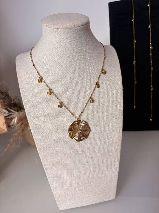 collier chaîne en acier inoxydable doré, orné d'un pendentif rond strié avec une pierre de lune et de pampilles rondes striées. Bijou présenté sur un buste en coton beige.