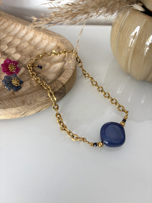 collier larges mailles en acier inoxydable doré, orné d'une grande perle losange bleu-gris scintillant. Bijou posé sur une table en verre trempée, décorée d'un bol en céramique kaki et un plateau en bambou sur lequel sont posées des boucles d'oreilles fleur hibiscus framboise et bleu nuit.