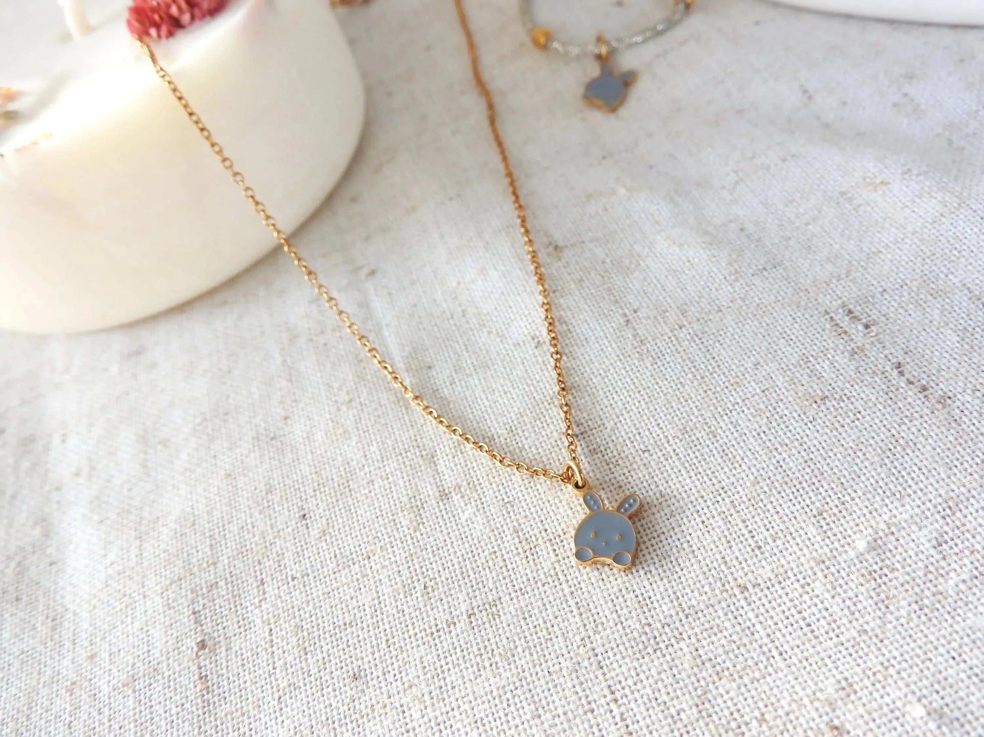 Collier enfant à chaîne fine en acier inoxydable doré et orné d'un pendentif lapin gris en résine époxy et acier inoxydable doré. Bijou posé à plat sur une nappe beige décorée d'une bogie fleurie.