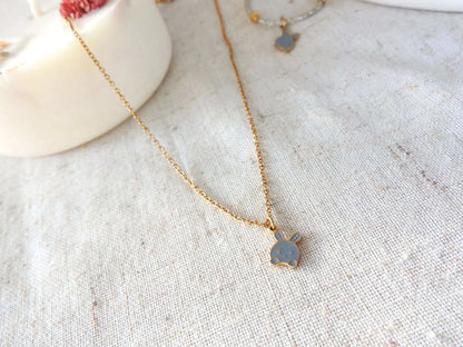 Collier enfant à chaîne fine en acier inoxydable doré et orné d'un pendentif lapin gris en résine époxy et acier inoxydable doré. Bijou posé à plat sur une nappe beige décorée d'une bogie fleurie.