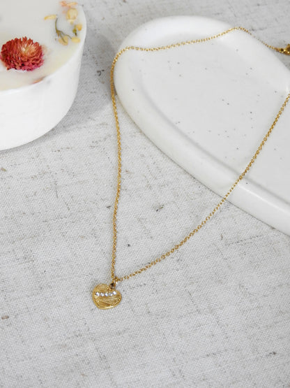Collier pendentif coeur texturé orné de pierres en oxydes de zirconium et en acier inoxydable doré. Bijou posé à plat sur une nappe beige en coton et coupelle en céramique blanche décorée d'une bougie fleurie.