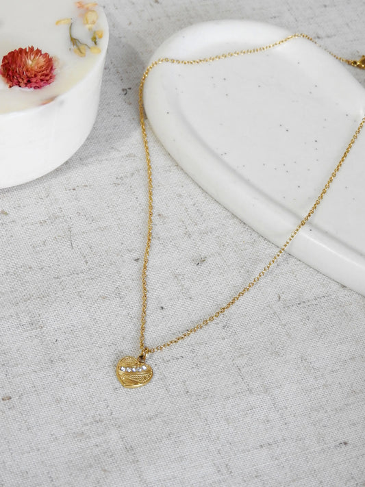 Collier pendentif coeur texturé orné de pierres en oxydes de zirconium et en acier inoxydable doré. Bijou posé à plat sur une nappe beige en coton et coupelle en céramique blanche décorée d'une bougie fleurie.