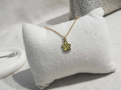 Collier pour enfant à chaîne fine posé sur un mini-coussin beige. Bijou orné d'un pendentif abeille jaune en résine époxy et acier inoxydable doré. 