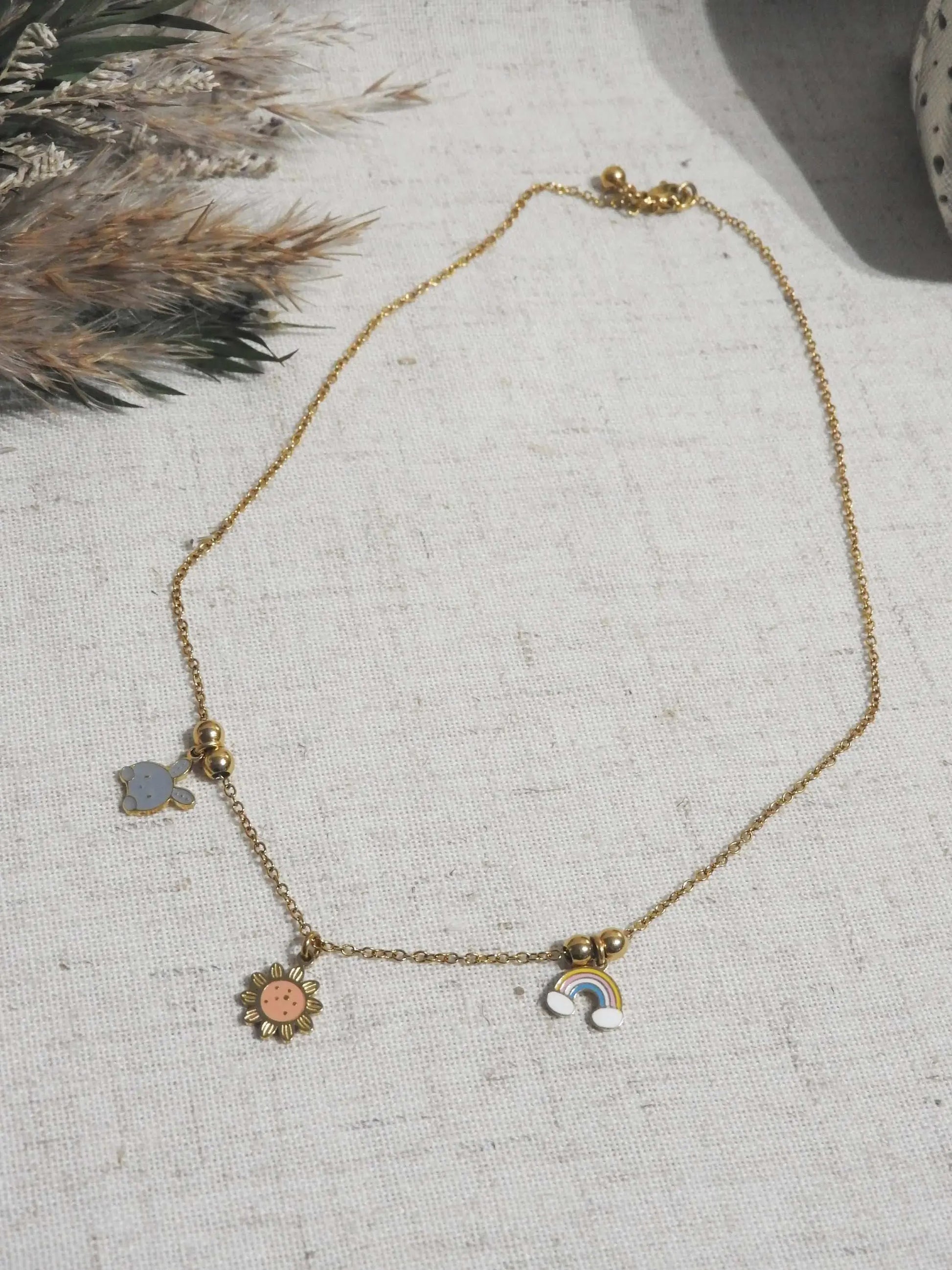 Collier enfant à chaîne fine en acier inoxydable doré et orné de trois pendentifs espacés par des petites perles dorées : lapin, arc-en-ciel et tournesol en résine époxy et acier inoxydable doré. Bijou posé à plat sur une nappe beige en coton, décorée d'un mini-bouquet de fleurs.