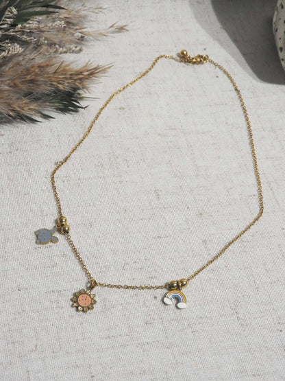 Collier enfant à chaîne fine en acier inoxydable doré et orné de trois pendentifs espacés par des petites perles dorées : lapin, arc-en-ciel et tournesol en résine époxy et acier inoxydable doré. Bijou posé à plat sur une nappe beige en coton, décorée d'un mini-bouquet de fleurs.