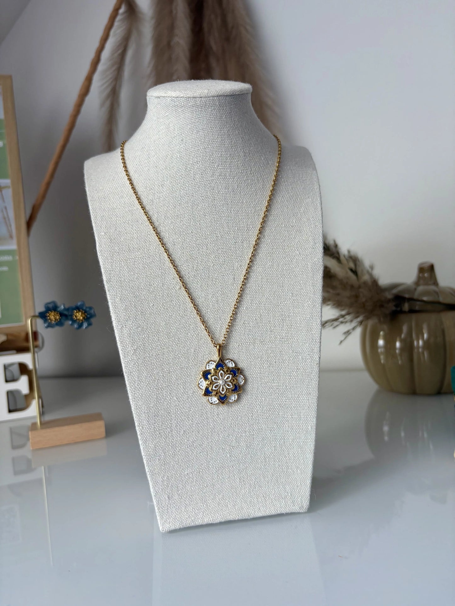 Collier sautoir en acier inoxydable doré présenté sur un buste présentoir. Bijou orné d'un pendentif mandala bleu et blanc en résine époxy et en acier inoxydable doré.