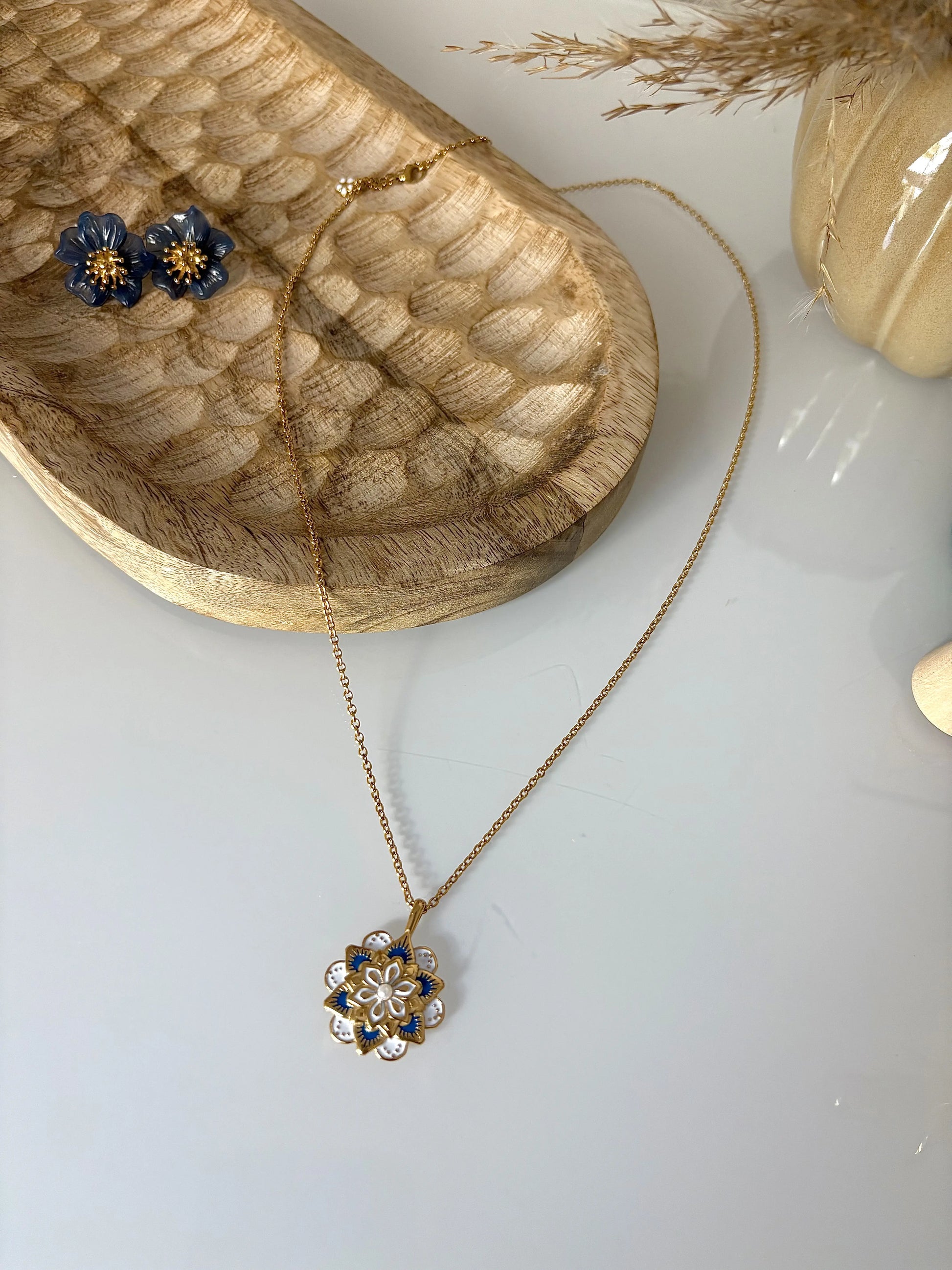 collier sautoir en acier inoxydable doré et orné d'un pendentif mandala bleu et blanc. Bijou posé sur un plateau en bambou sur lequel sont disposées des boucles d'oreilles fleurs bleu foncé.