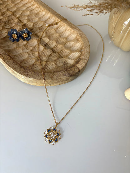 collier sautoir en acier inoxydable doré et orné d'un pendentif mandala bleu et blanc. Bijou posé sur un plateau en bambou sur lequel sont disposées des boucles d'oreilles fleurs bleu foncé.