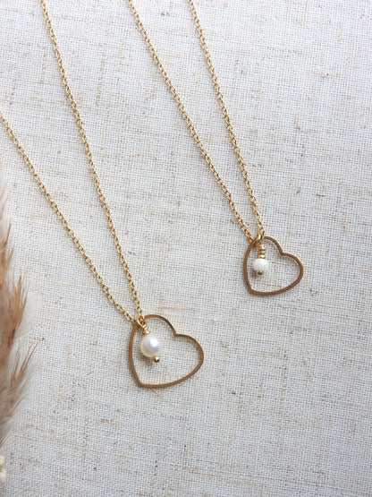 deux colliers côte à côte posés à plat sur une nappe beige, chaîne fine en acier inoxydable doré avec un pendentif coeur évidé et orné l'un avec une perle d'eau douce et une perle de verre pour le 2ème collier.
