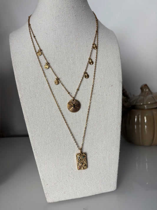 Colliers multi-rangs en acier inoxydable doré et ornés de deux pendentifs, l'un rectangle avec symboles et l'autre rond strié. Bijou présenté sur un buste en coton beige disposé sur une table.