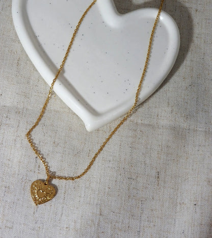 Collier chaîne pendentif coeur gravé de deux mains supportant un coeur. Bijou en acier inoxydable doré posé à plat sur une nappe beige et une coupelle en céramique blanche.