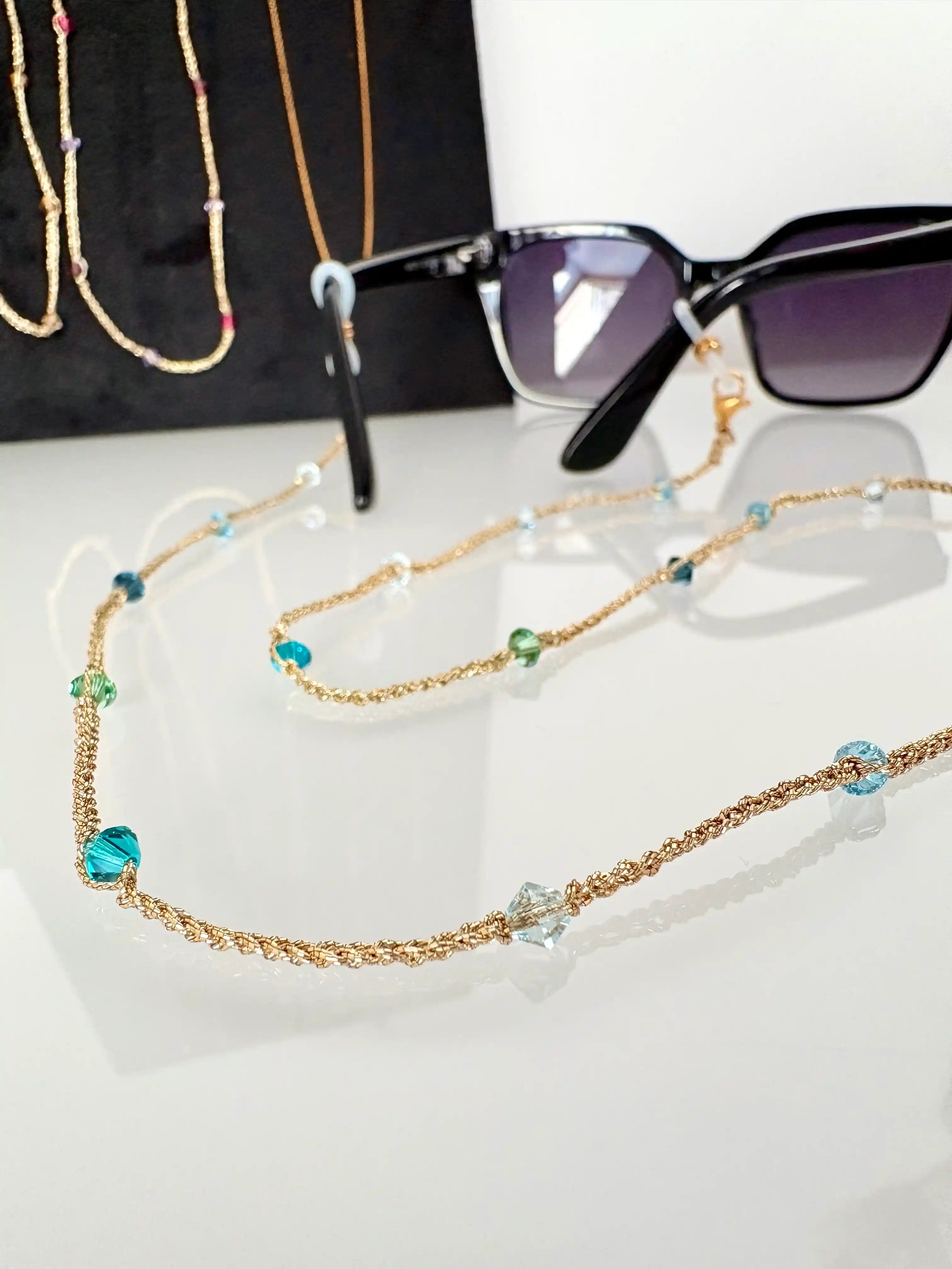 Bijou cordon à lunettes à fil doré crocheté à la main. Bijou orné de perles toupies en cristal aux nuances bleu et vert. Bijou monté sur monture de lunettes de soleil noire.