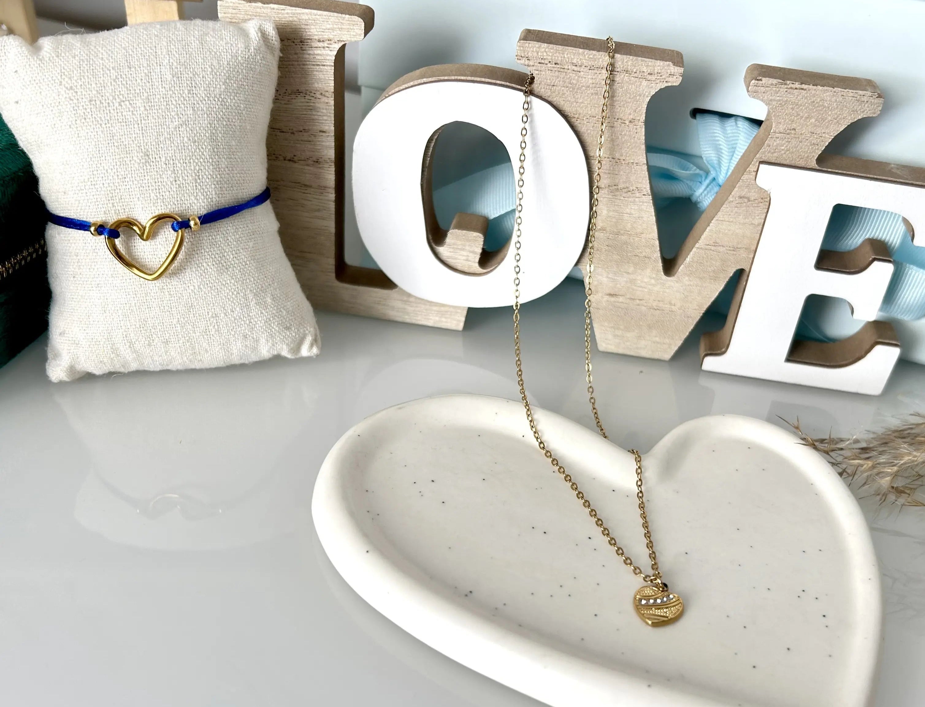 décoration d'une table en verre trempée avec l'inscription LOVE en bois, avec une coupelle coeur en céramique blanche, sur laquelle est posée un collier chaîne fine avec un pendentif coeur orné de pierres en oxyde de zirconium et acier doré ainsi qu'un bracelet cordon bleu roi avec un coeur intercalaire en acier inoxydable doré.