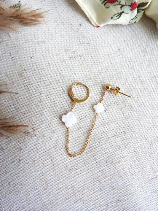 Double boucle d'oreilles à clou boule et une mini-créole reliée par une chainette ornée de deux perles trèfle en nacre blanche et en acier inoxydable doré. Bijou posé sur une nappe beige décorée d'une brindille.