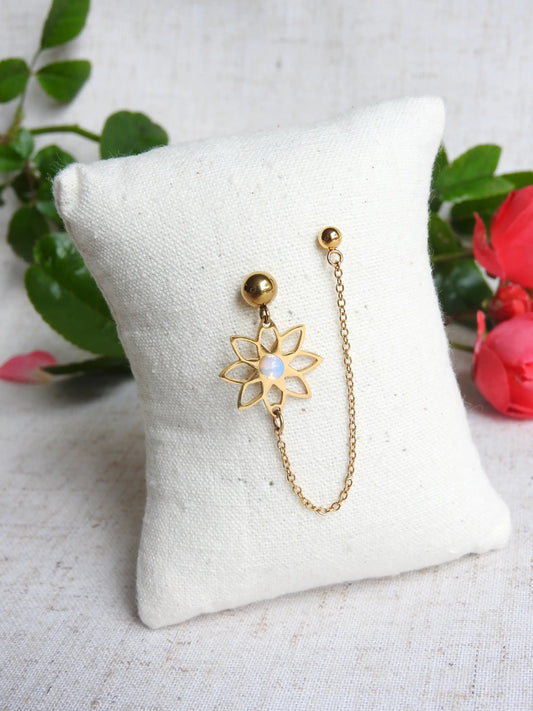 double boucles d'oreilles clous à boule reliées par une chaînette décorée d'une fleur de tournesol évidée et ornée d'une pierre en cristal. Bijou piqué sur un mini-coussin, posé sur une nappe beige décorée de roses.