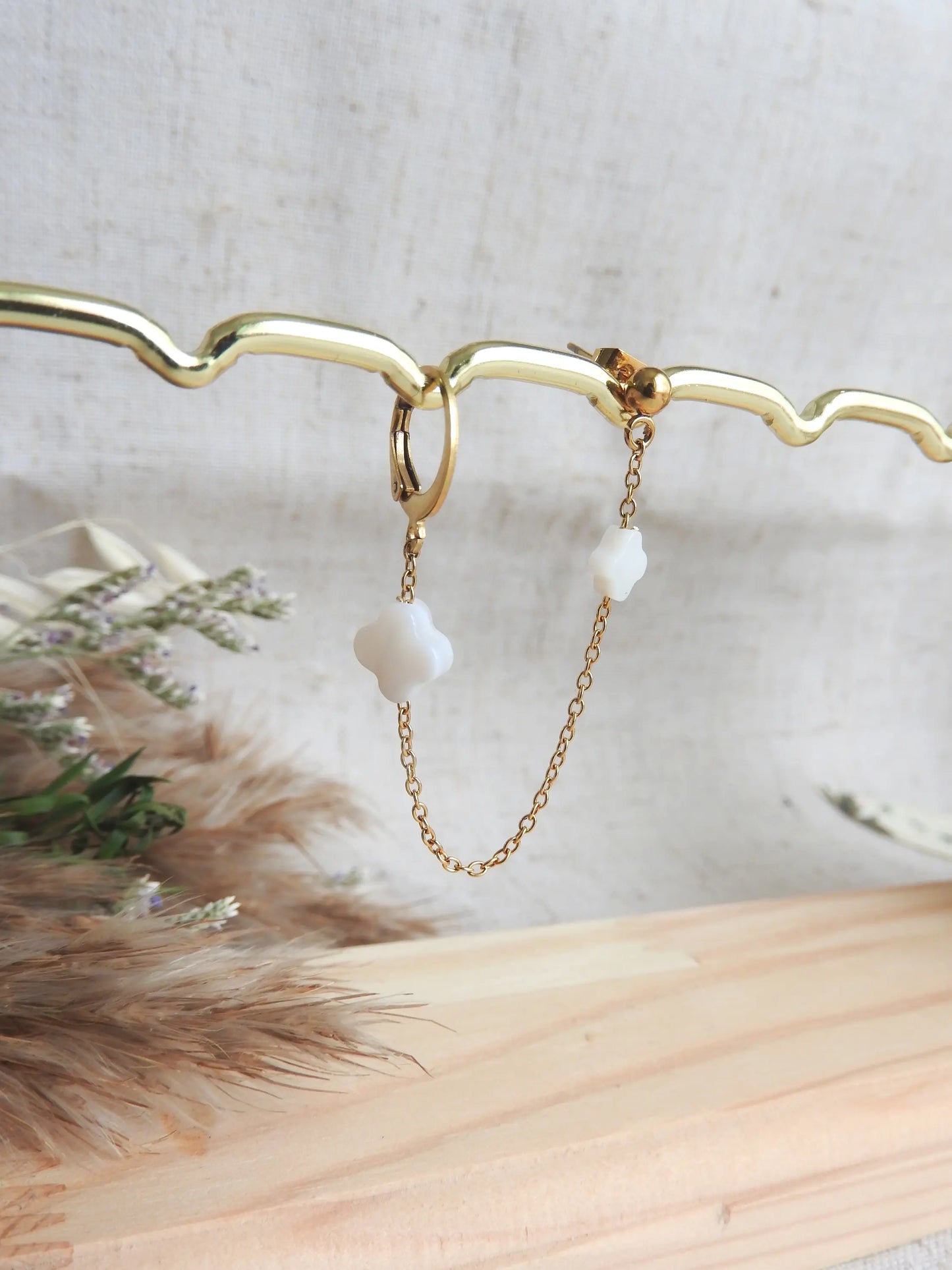 Double boucle d'oreilles à clou boule et une mini-créole reliée par une chainette ornée de deux perles trèfle en nacre blanche et en acier inoxydable doré. Bijou présenté sur une barre métallique doré et en bambou.