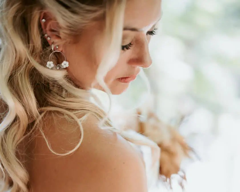 Mariée en tenue de mariage de profil et portant des boucles d'oreilles Jasmine de la marque de bijoux M'Joy Creation. Bijou créoles décorées de fleur blanche en nacre et strass.