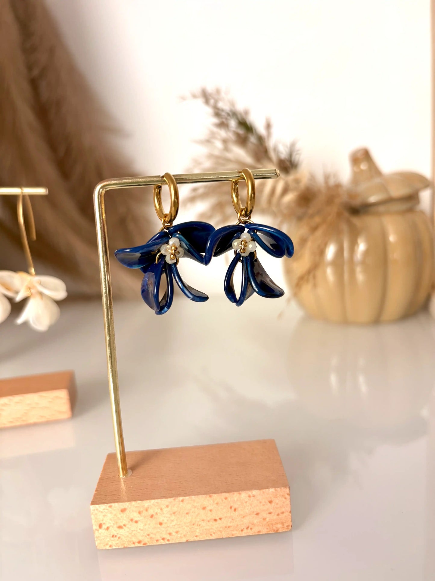 Boucles d'oreilles mini-créoles en acier inoxydable doré, ornées de pétales de fleurs en acrylique bleu roi marbré et perles de verre jaune pâle. Bijou présenté sur un support métallique doré et bambou posé sur une table en verre et décorée d'une citrouille décorative en céramique.