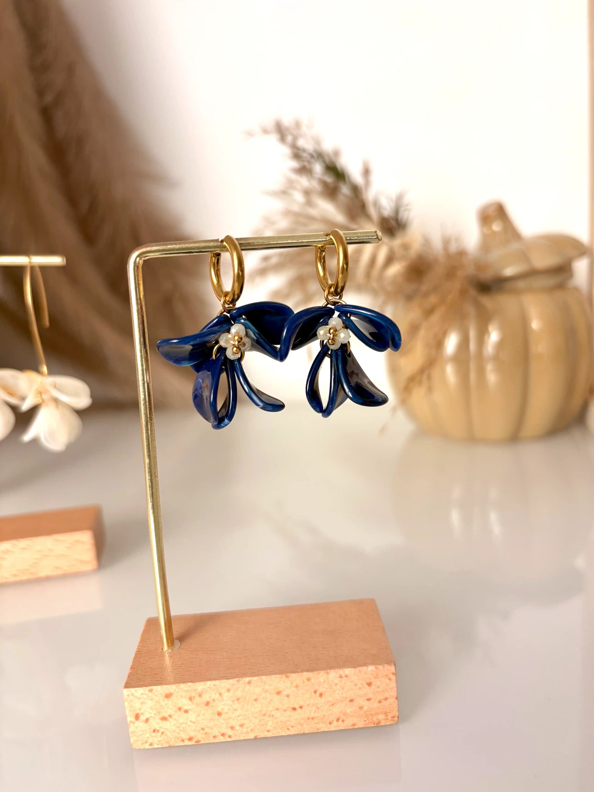 Boucles d'oreilles mini-créoles en acier inoxydable doré, ornées de pétales de fleurs en acrylique bleu roi marbré et perles de verre jaune pâle. Bijou présenté sur un support métallique doré et bambou posé sur une table en verre et décorée d'une citrouille décorative en céramique.