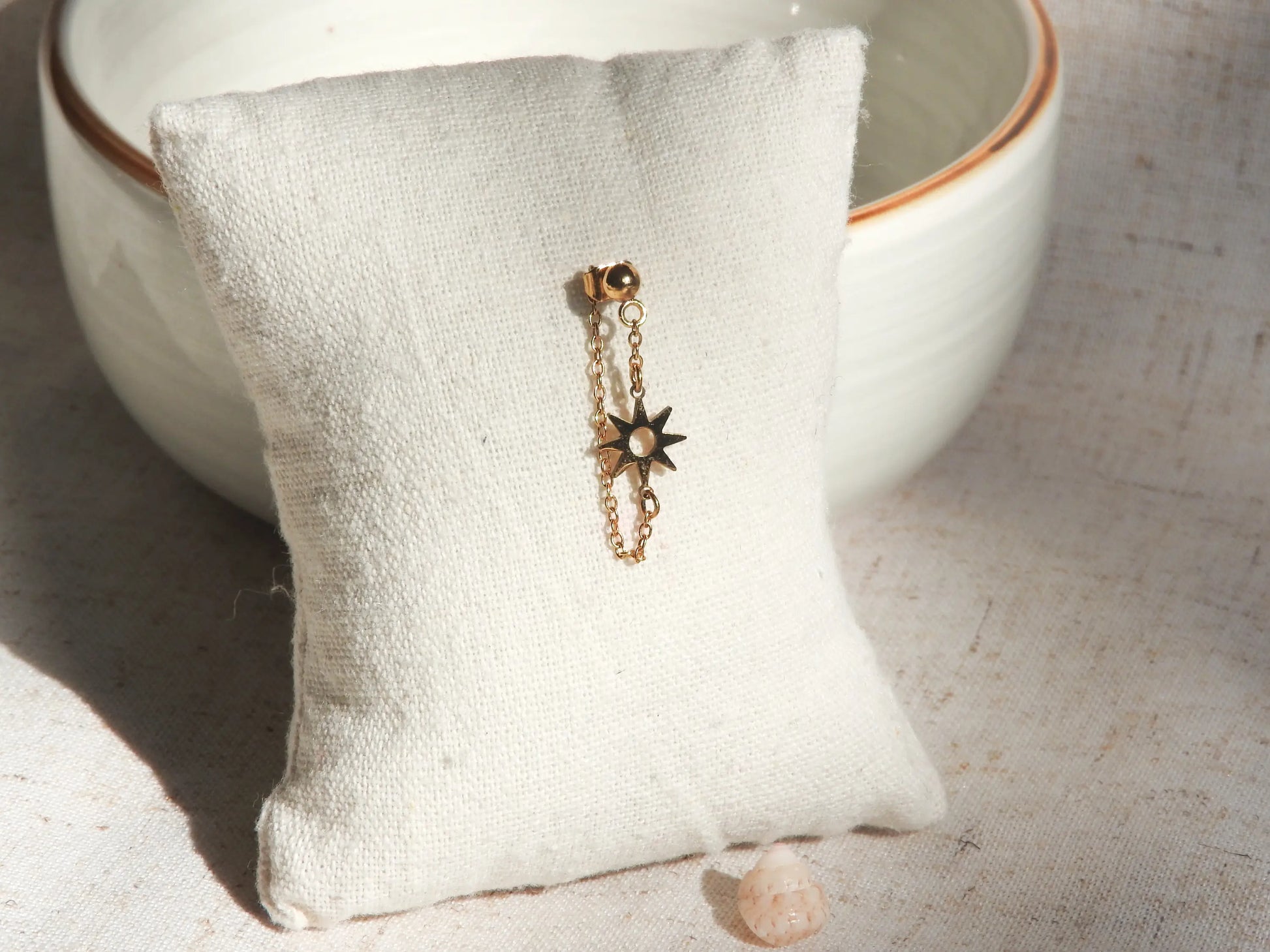 Mono-boucle d'oreille chaînette orné d'un soleil en intercalaire en acier inoxydable doré, présenté sur un mini-coussin beige en coton, posé contre un bol en céramique.