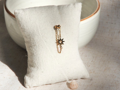Mono-boucle d'oreille chaînette orné d'un soleil en intercalaire en acier inoxydable doré, présenté sur un mini-coussin beige en coton, posé contre un bol en céramique.