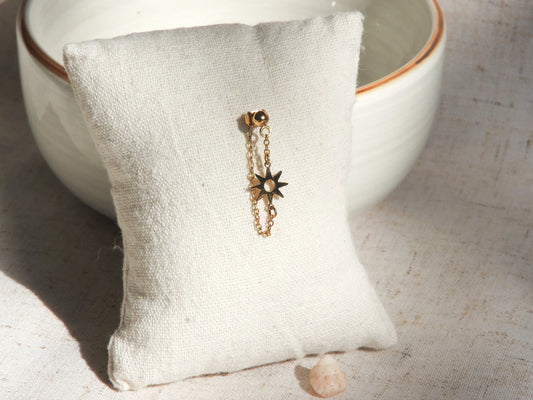 Mono-boucle d'oreille chaînette orné d'un soleil en intercalaire en acier inoxydable doré, présenté sur un mini-coussin beige en coton, posé contre un bol en céramique.