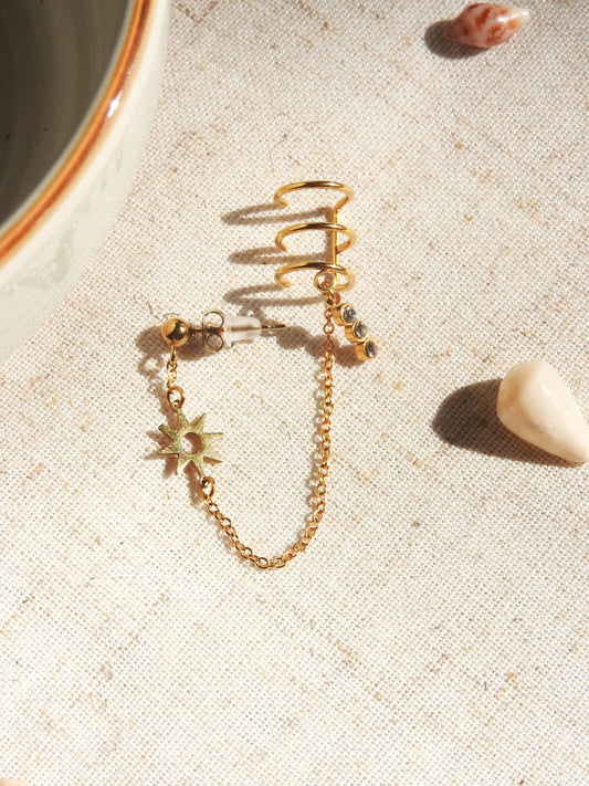 mono-boucle d'oreille clou boule chaînette reliée à une triple bague d'oreille en acier inoxydable doré. Bijou orné d'un soleil en intercalaire et une barre 3 pierres en oxydes de zirconium. Bijou posé à plat sur une nappe beige décorée de coquillages