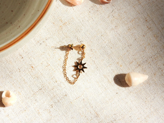Mono-boucle d'oreille à clou boule à chaînette en acier inoxydable doré, ornée d'un soleil en intercalaire. Bijou posé à plat sur une nappe beige décoré de coquillages.